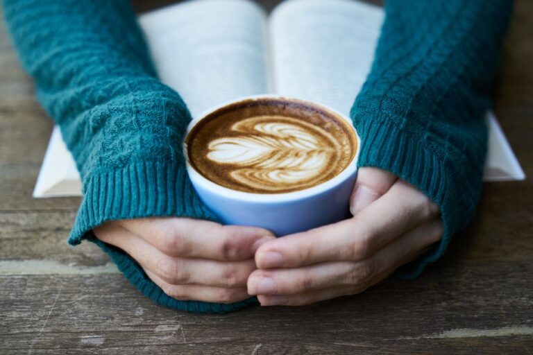 Hands holding a latte art coffee cup over an open book on a wooden table.