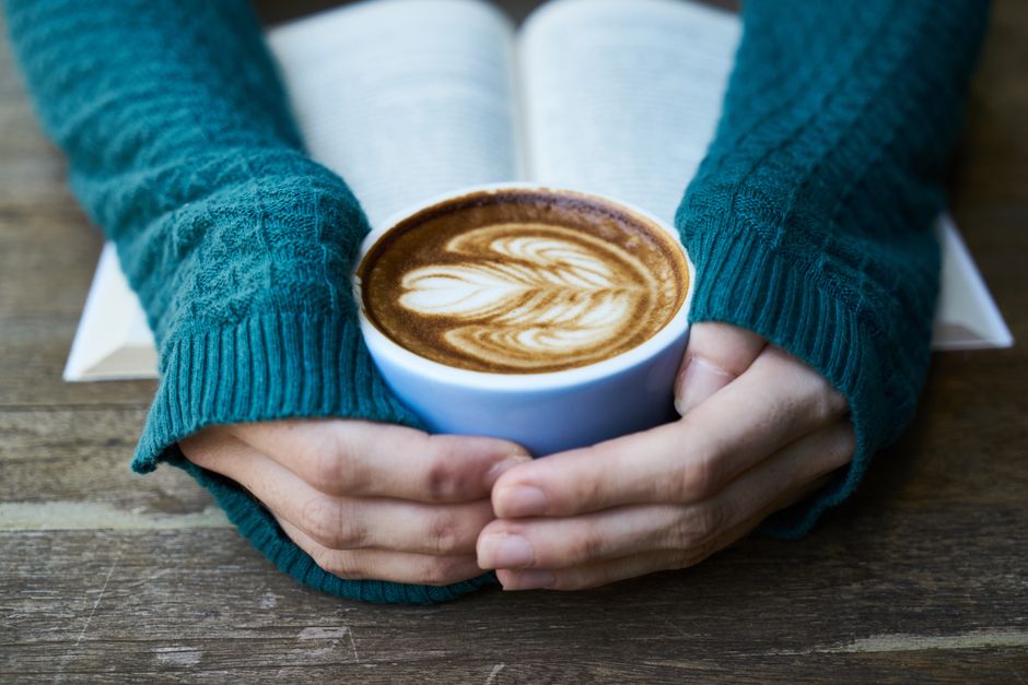 Hands holding a latte art coffee cup over an open book on a wooden table.