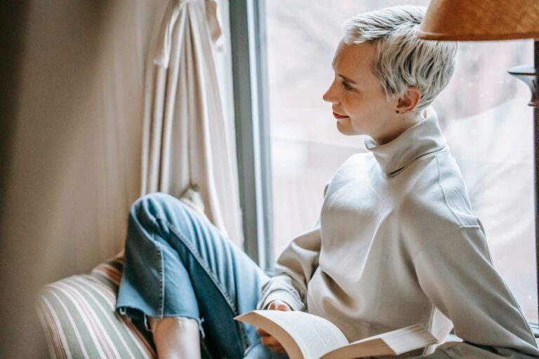 Side view of clever female with stylish haircut relaxing with book on window in daytime