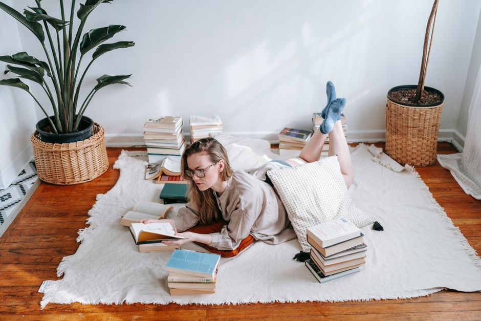 Concentrated young female student with long wavy blond hair in casual sweater and eyeglasses reading book with interest while sitting near green potted plant in light apartment